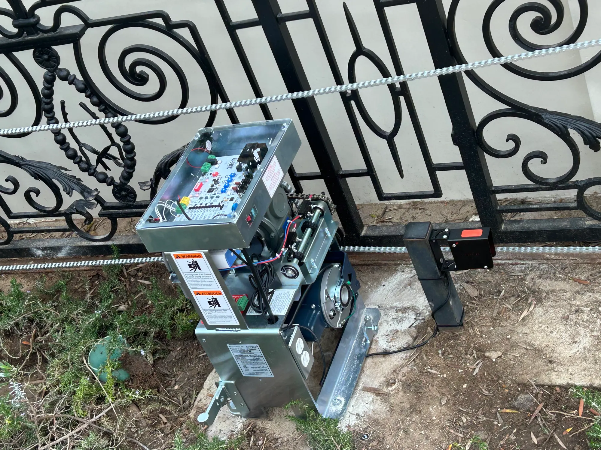 An All-O-Matic slide gate operator with its control enclosure open beside an ornamental iron gate, showing the board, wiring and drive rack