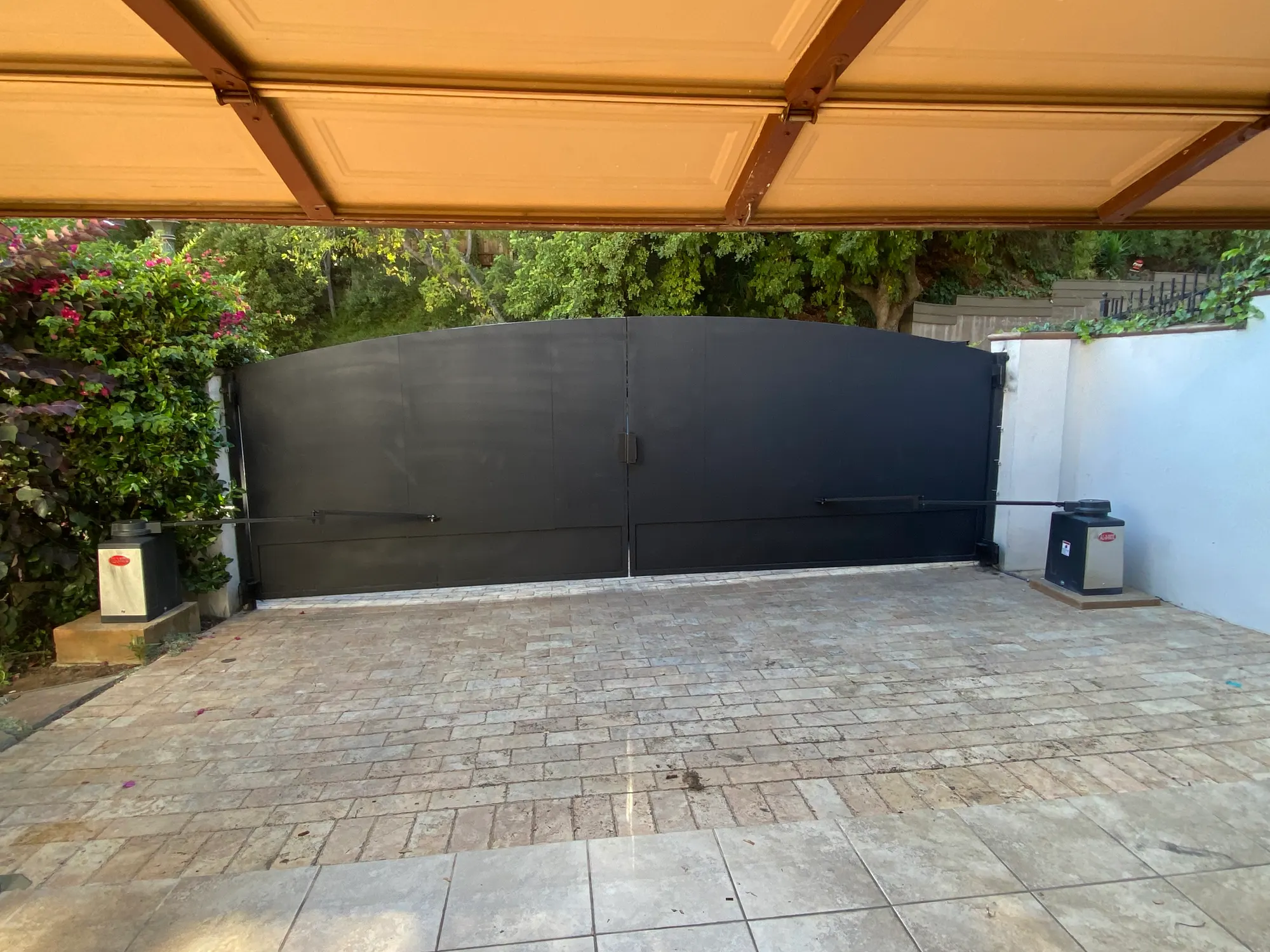 A pair of tall dark metal swing gates closed across a paved driveway beneath a timber carport