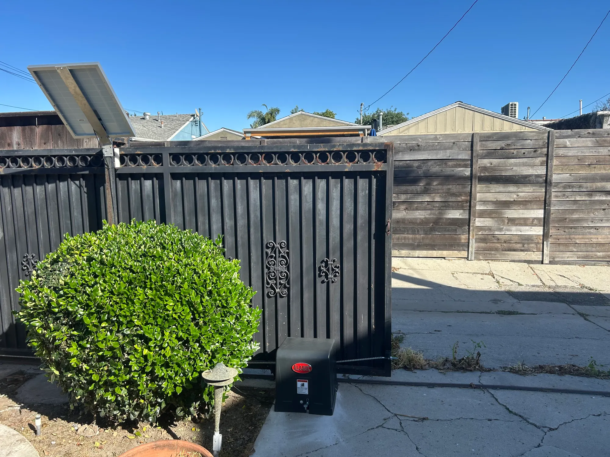 A black slide gate operator mounted on a concrete pad beside a decorative iron gate and clipped hedge