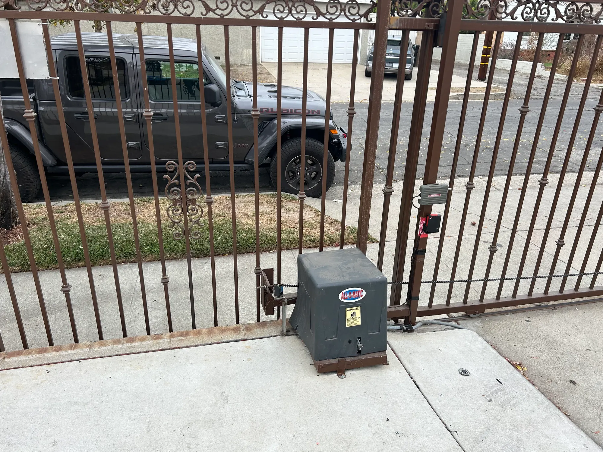 A gate operator in a grey housing installed on a concrete pad at the base of a tall iron gate, with the drive arm connected to the gate frame