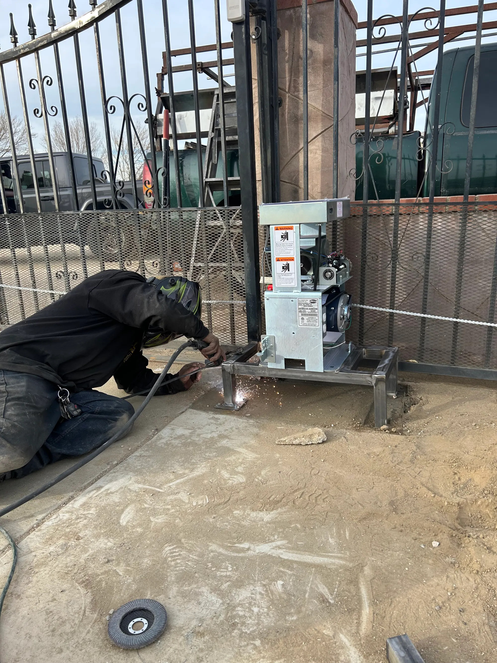 A technician kneeling at a gate operator base, welding the mounting frame in place during installation