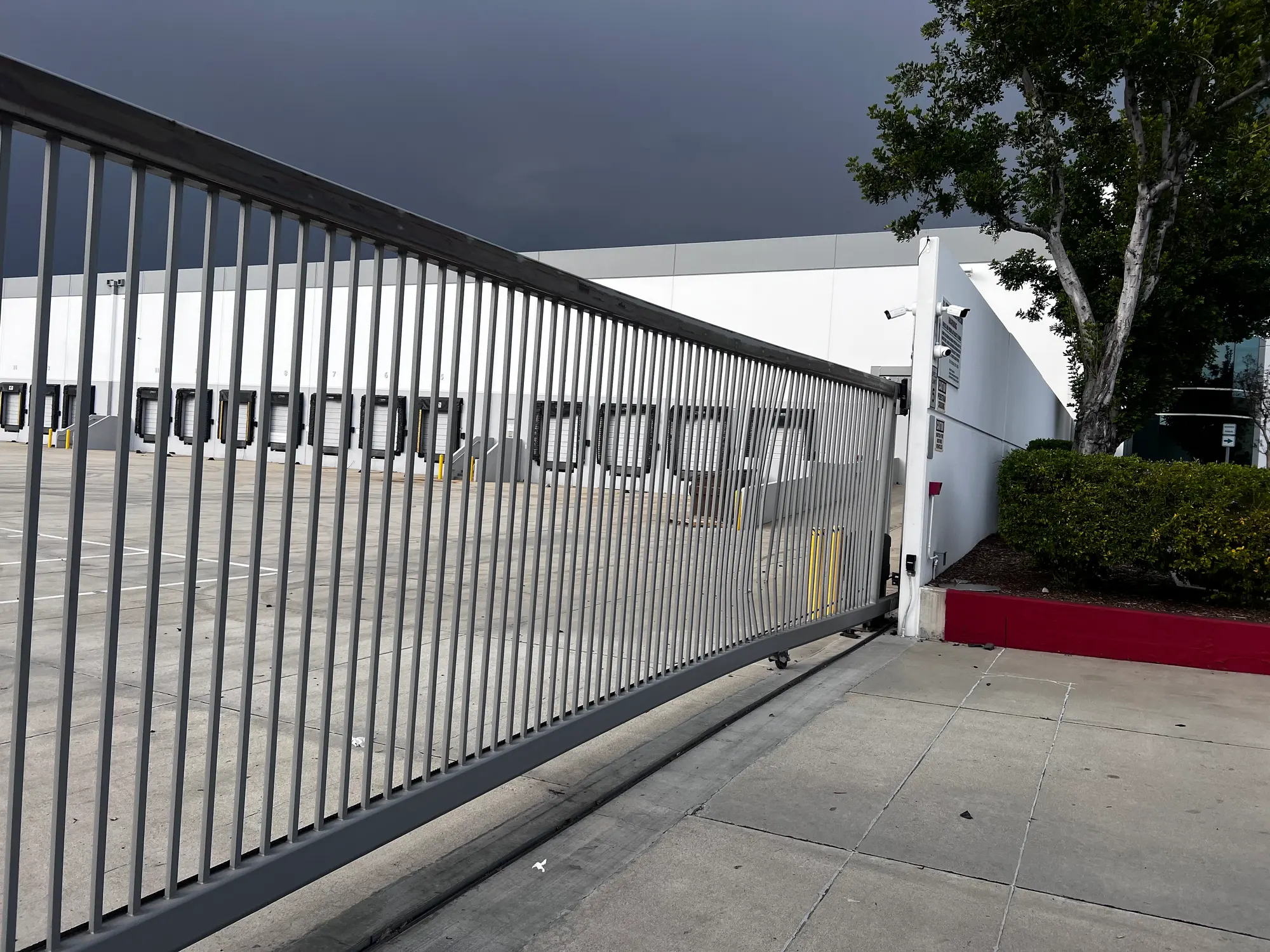 A long steel picket slide gate closed across the entrance to a distribution warehouse, with loading dock doors behind it