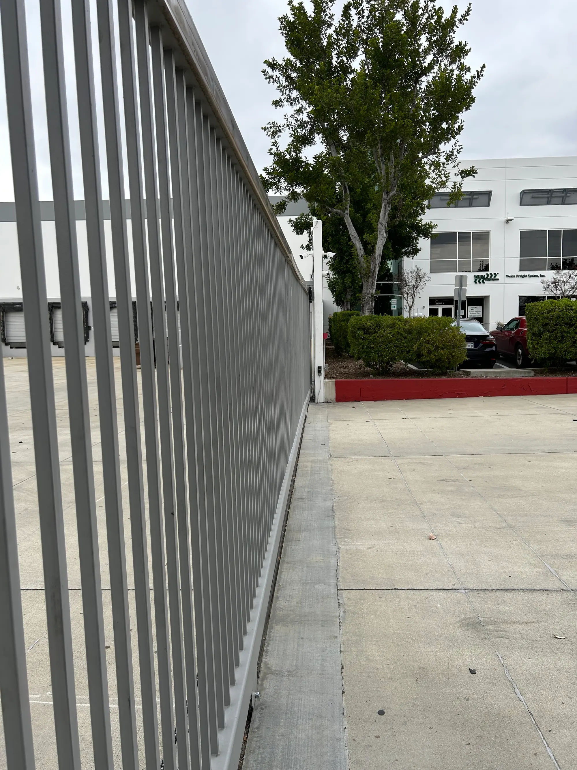 A long view down the repaired steel picket gate standing closed and square on its track, with the office building and yard beyond