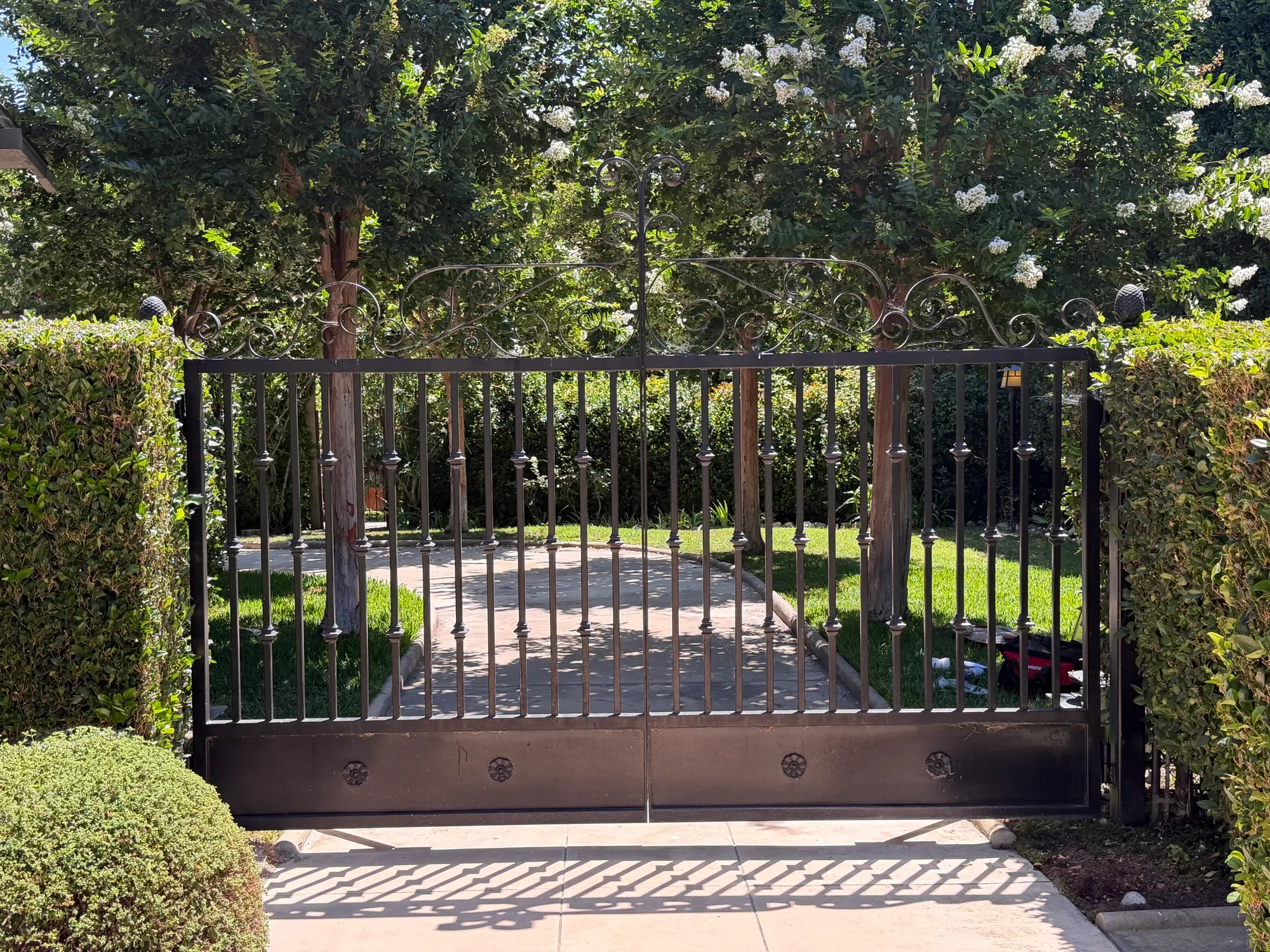A pair of black iron swing gates closed across a garden entrance, with planting and a paved path behind them