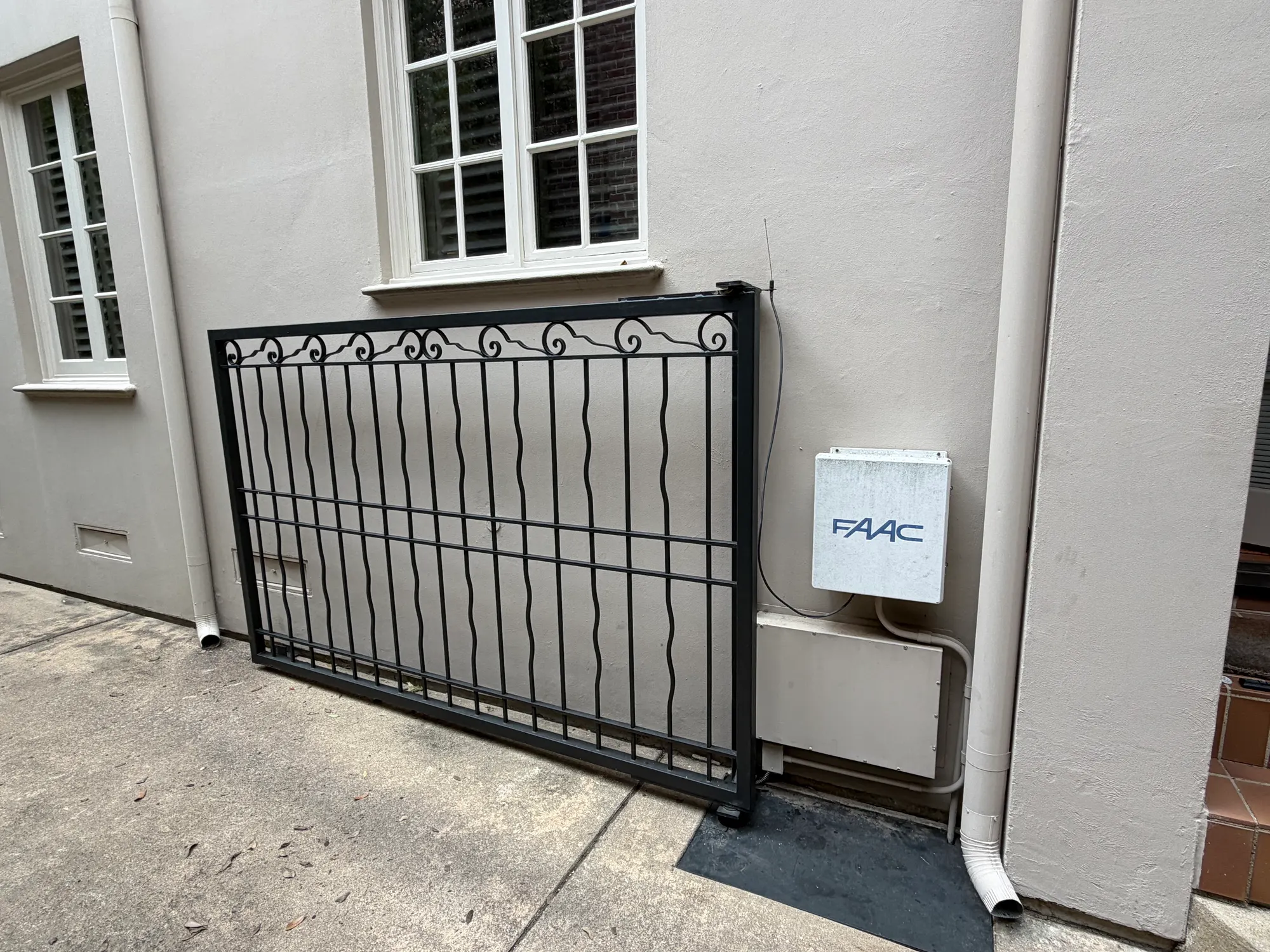 A black wrought-iron slide gate closed alongside a rendered wall, with a FAAC control enclosure mounted on the wall beside it and its aerial running above