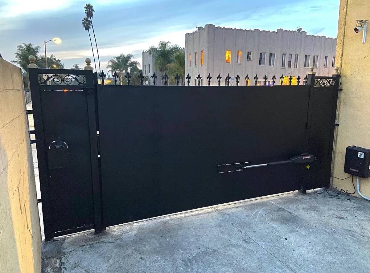 A tall black solid-panel swing gate closed across a driveway entrance, with a LiftMaster swing operator arm mounted along the lower rail and a control box on the wall beside it
