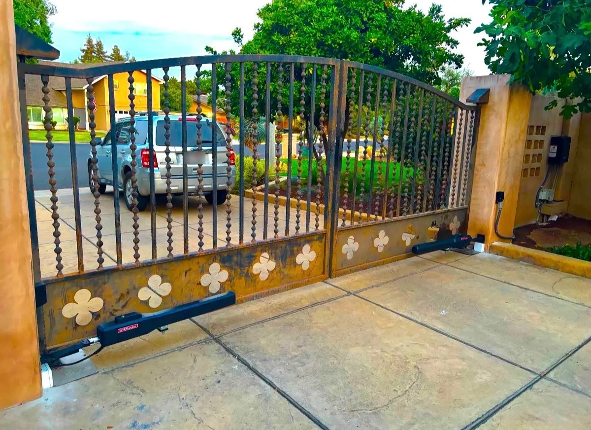 A decorative wrought-iron double driveway gate with ornamental scrollwork, fitted with a LiftMaster operator arm at the base of each leaf