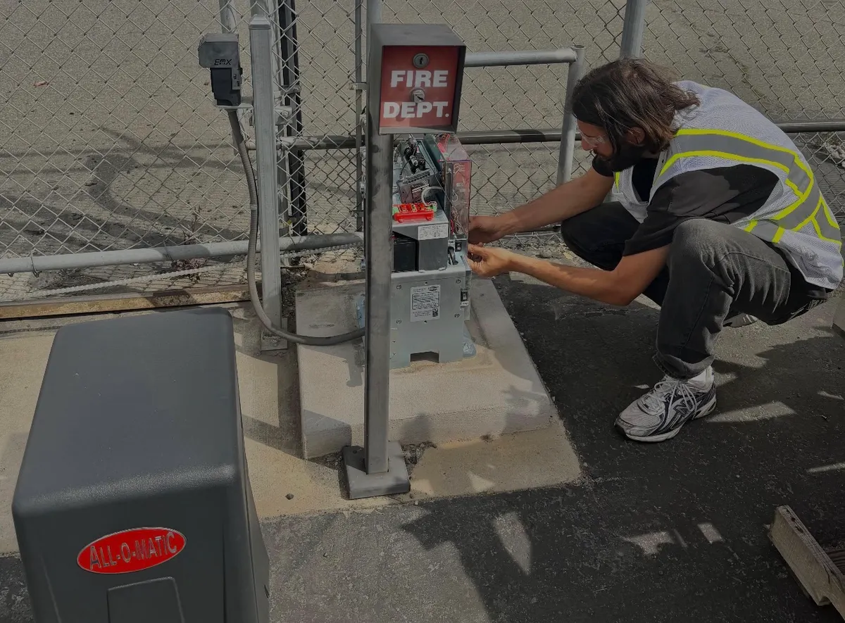 A Shield Gate Repair technician in a high-visibility vest crouched at an opened gate control pedestal, testing the wiring terminals by hand, with an All-O-Matic slide operator in the foreground
