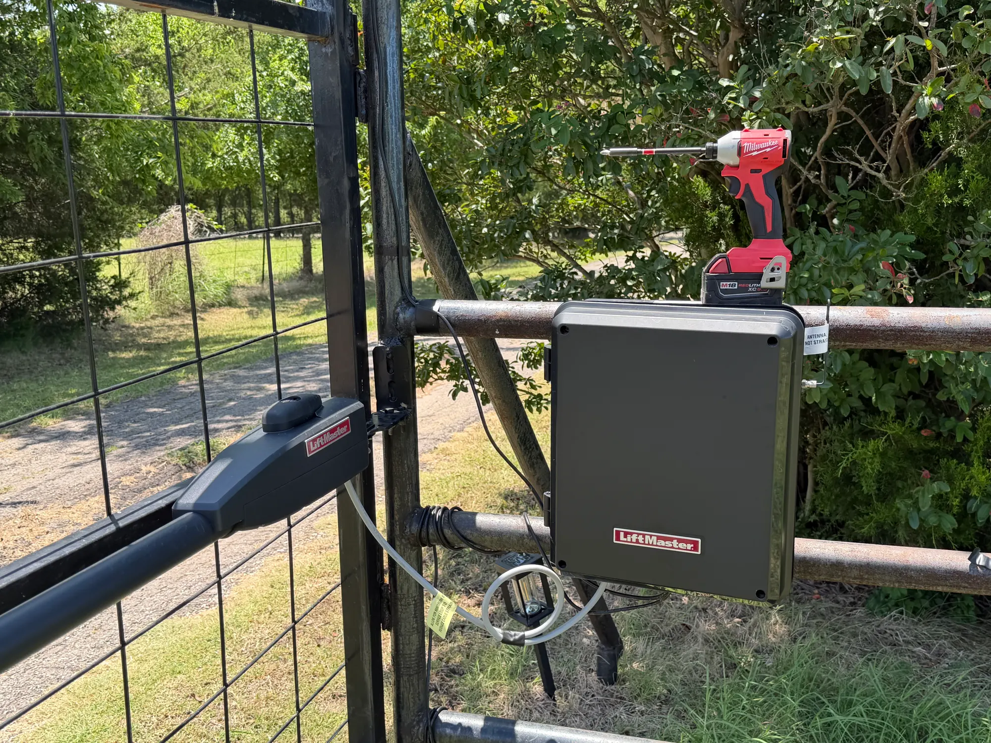 A LiftMaster control enclosure mounted on the post of a rural wire-mesh gate, with an impact driver resting on top of the housing during the repair