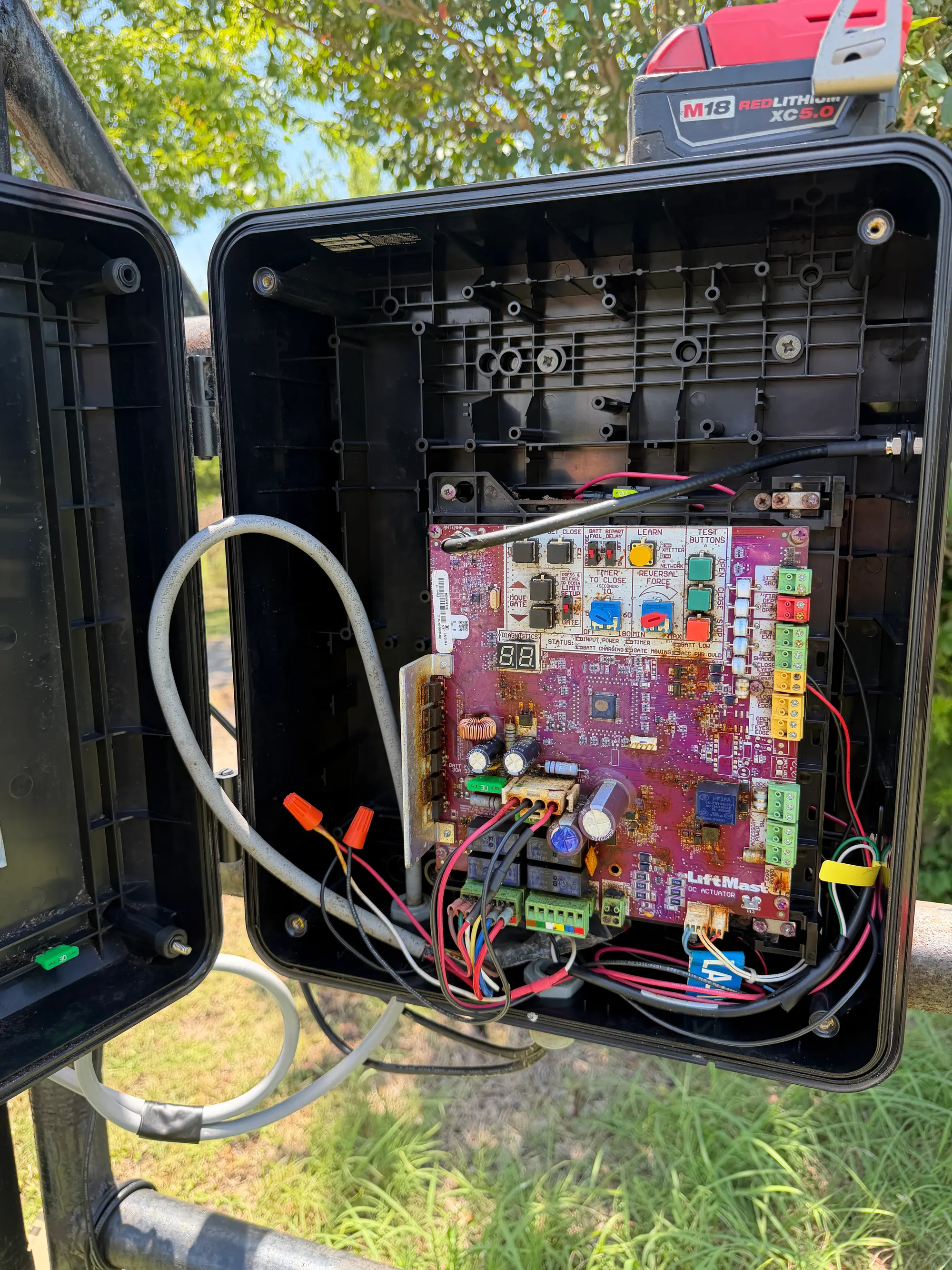 A LiftMaster gate operator enclosure standing open beside the gate, showing the control board and wiring loom inside