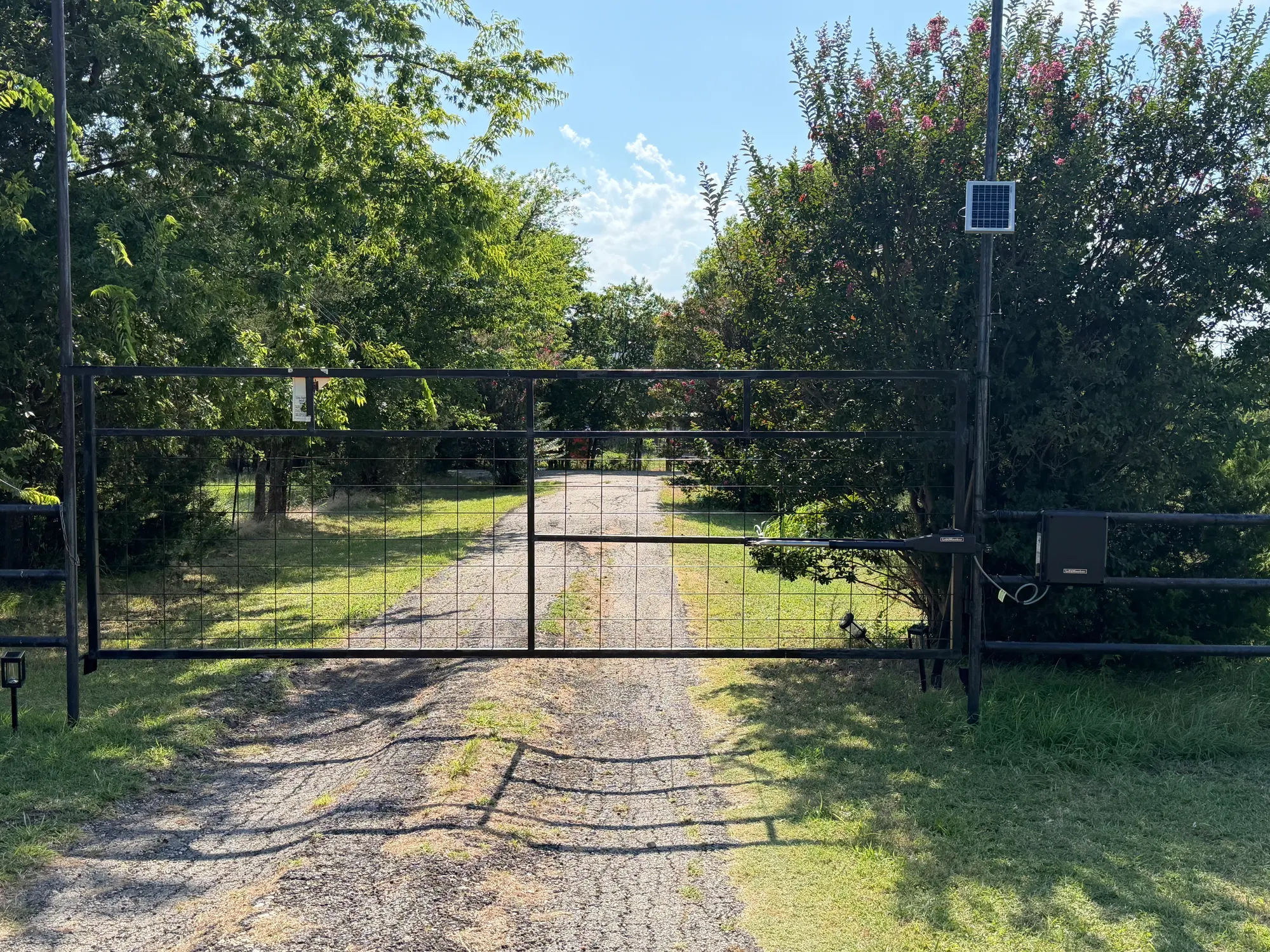 A double farm gate closed across a long tree-lined driveway, with a solar panel mounted on the post to the right of the gate