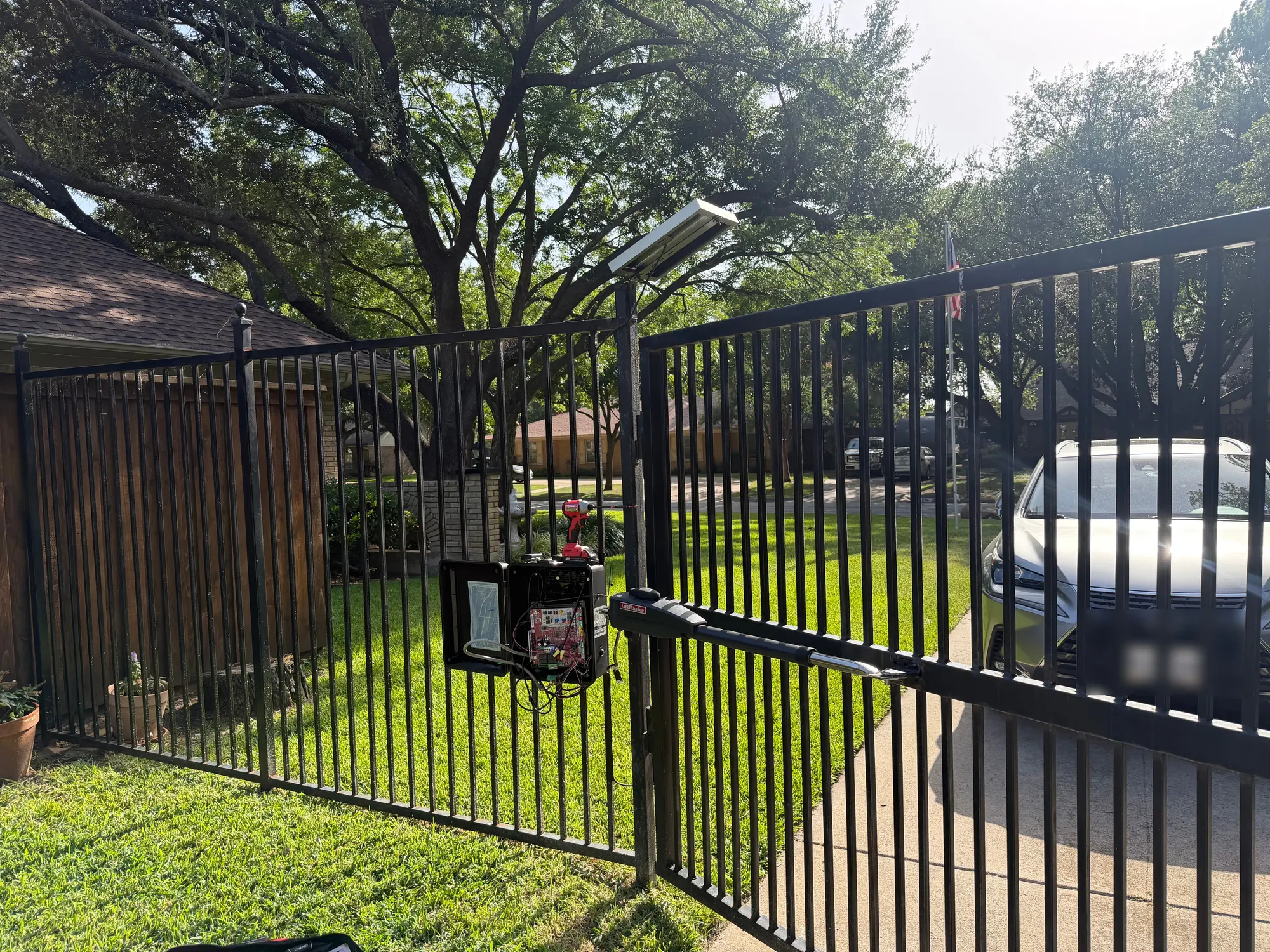 A LiftMaster swing gate operator on a black iron driveway gate, with the control enclosure open and the circuit board exposed mid-diagnosis, an impact driver resting on the housing and a solar panel mounted above on the gate post