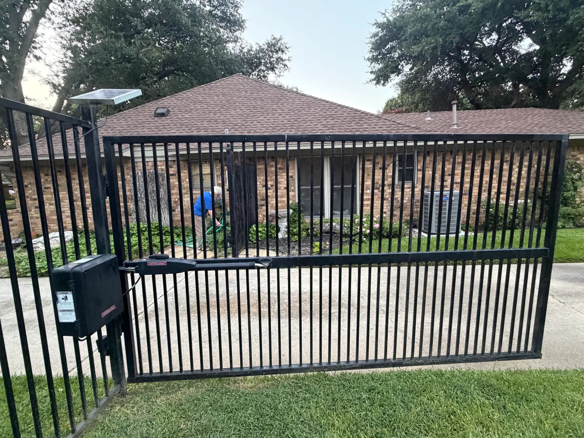 The completed job — a black iron driveway gate closed across a suburban brick home, with the LiftMaster operator, sealed control box and post-mounted solar panel all back in service