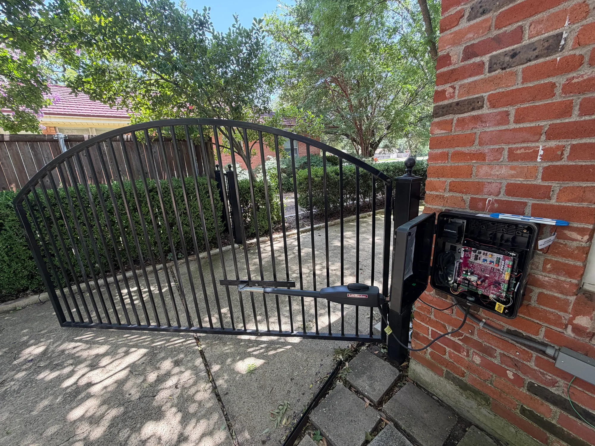 An arched wrought iron swing gate standing open on a residential walkway, with its LiftMaster linear arm operator attached to the gate frame and the wall-mounted control box open on the brick pier alongside