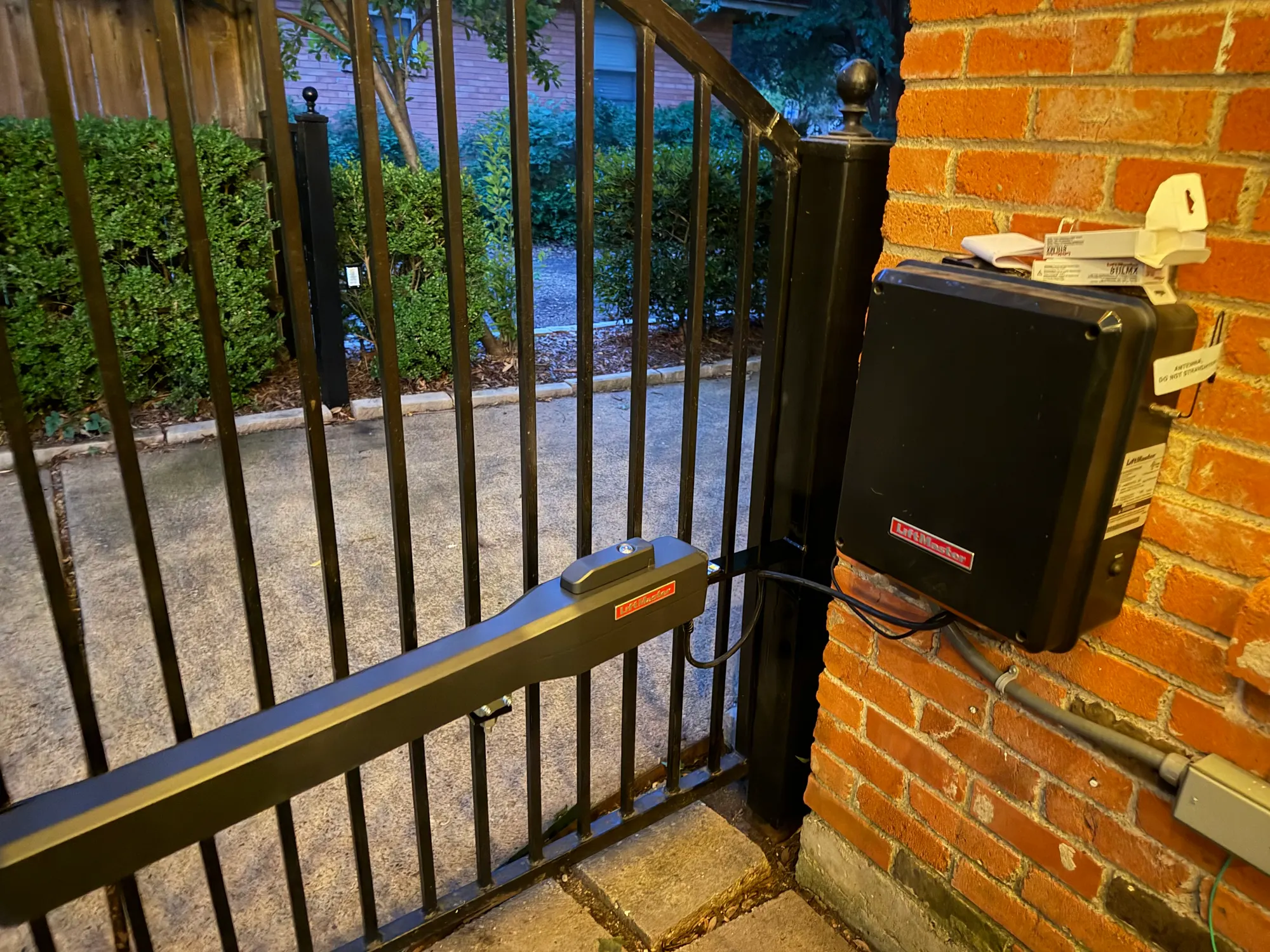 The upgraded LiftMaster arm fitted to the iron gate at dusk, with the original control box closed and still mounted on the same brick pier and conduit run