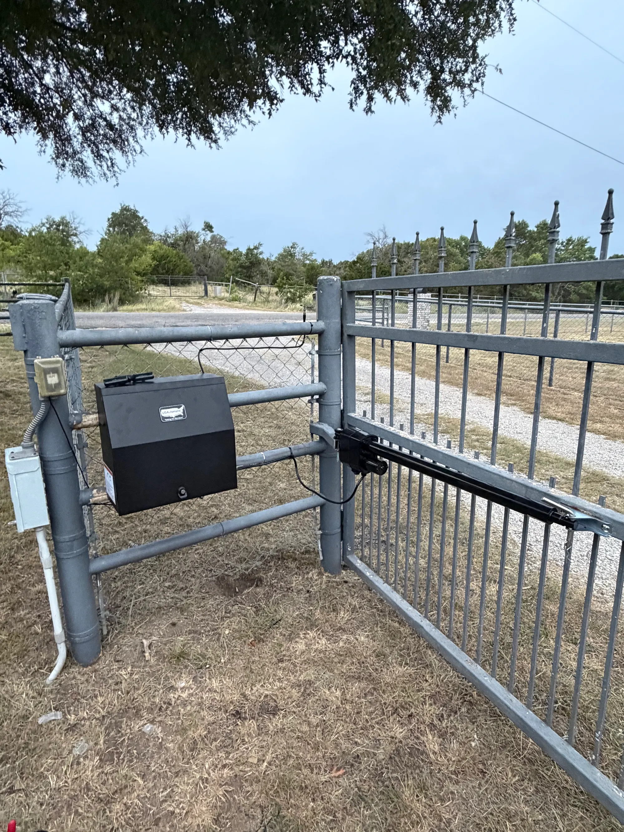 A gate operator in a black weatherproof enclosure mounted on the post of a metal ranch gate, with the drive arm connected to the gate frame