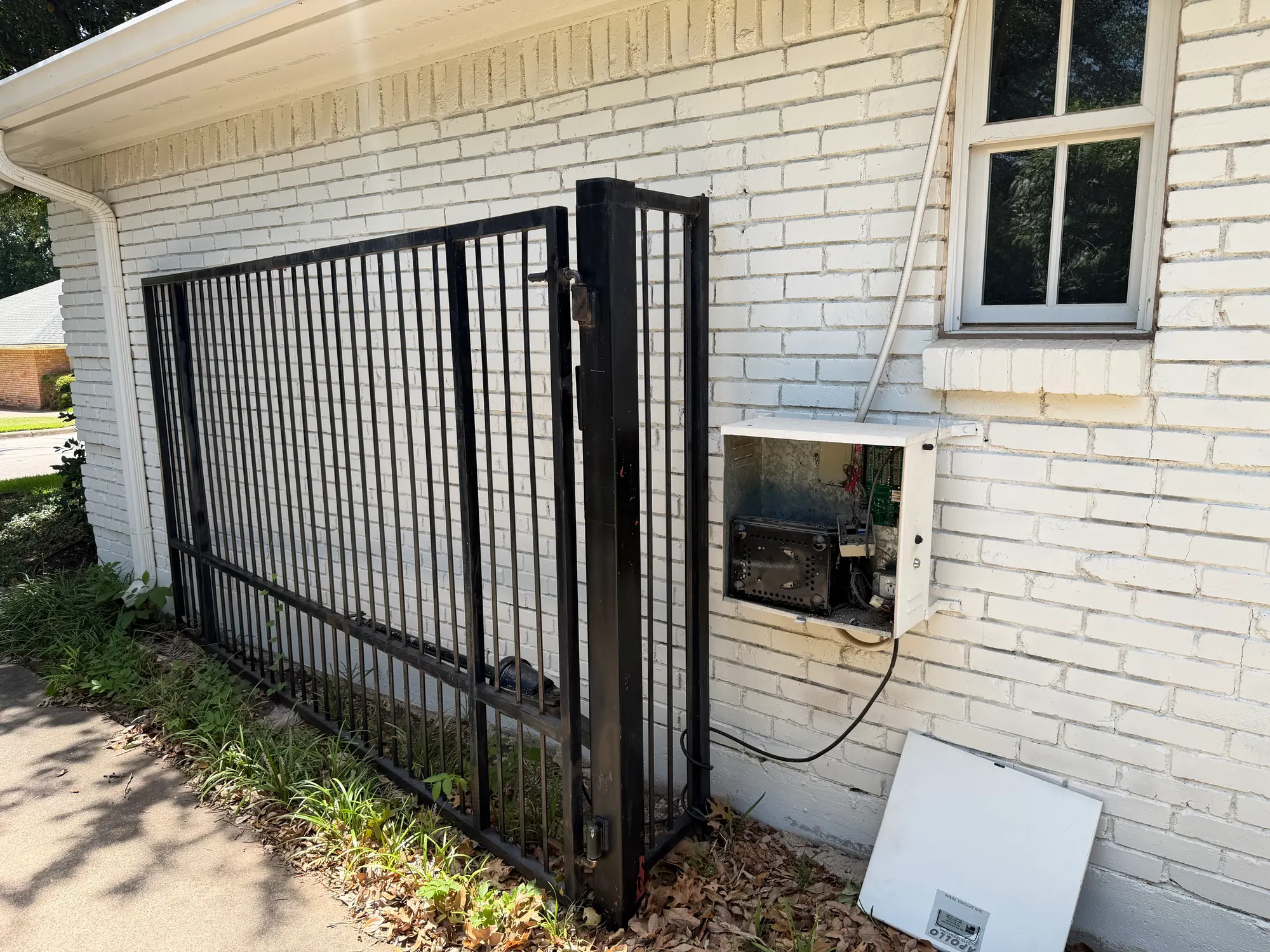 A black metal gate beside a brick wall with its control enclosure open, showing the operator wiring and components inside