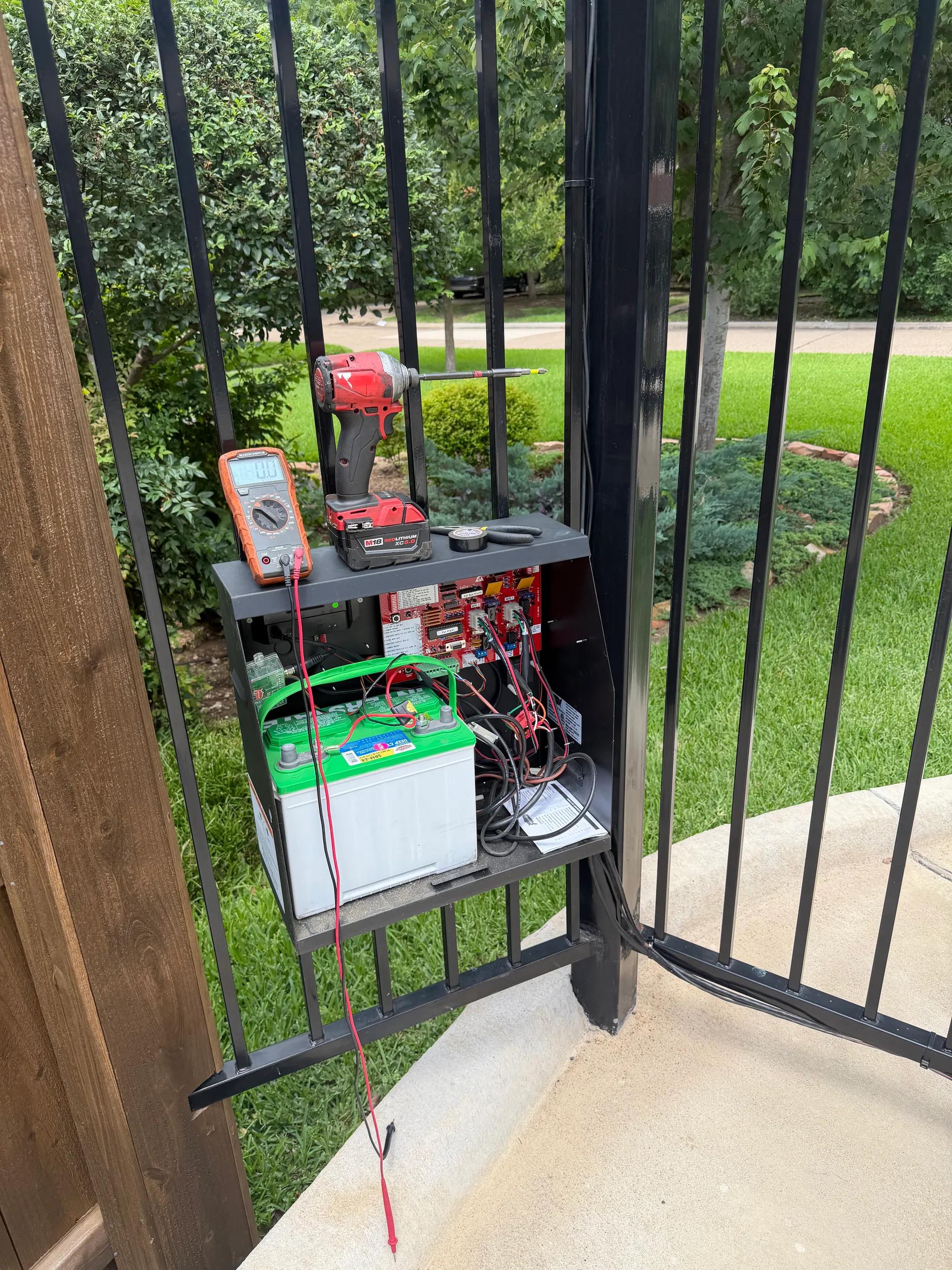 An opened gate control enclosure showing the battery, charge wiring and control board, with a multimeter resting on top during electrical testing