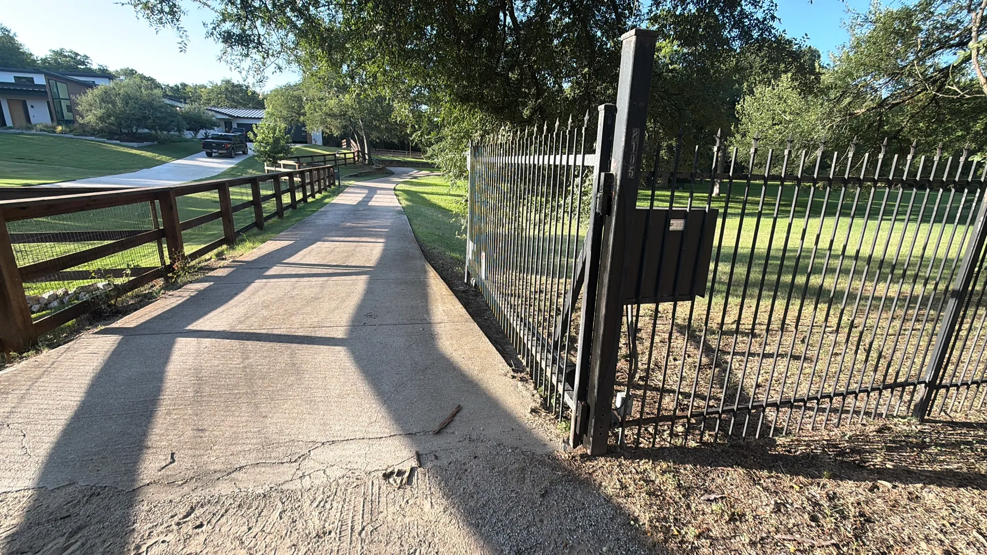 A metal gate at the entrance to a long tree-lined driveway, with the operator control box mounted on the right-hand post